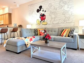 Un salon avec un canapé gris, deux tabourets de bar, une table basse et une fresque murale représentant Mickey Mouse.