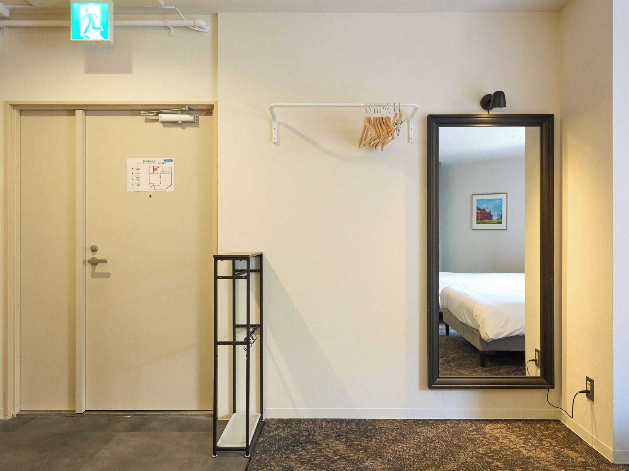 Une chambre d’hôtel avec un lit, un grand miroir, une petite table et une porte portant un panneau indiquant la sortie de secours.