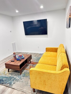 Un salon avec un canapé jaune, une table basse en bois, un téléviseur à écran plat fixé au mur et un tapis à motifs.