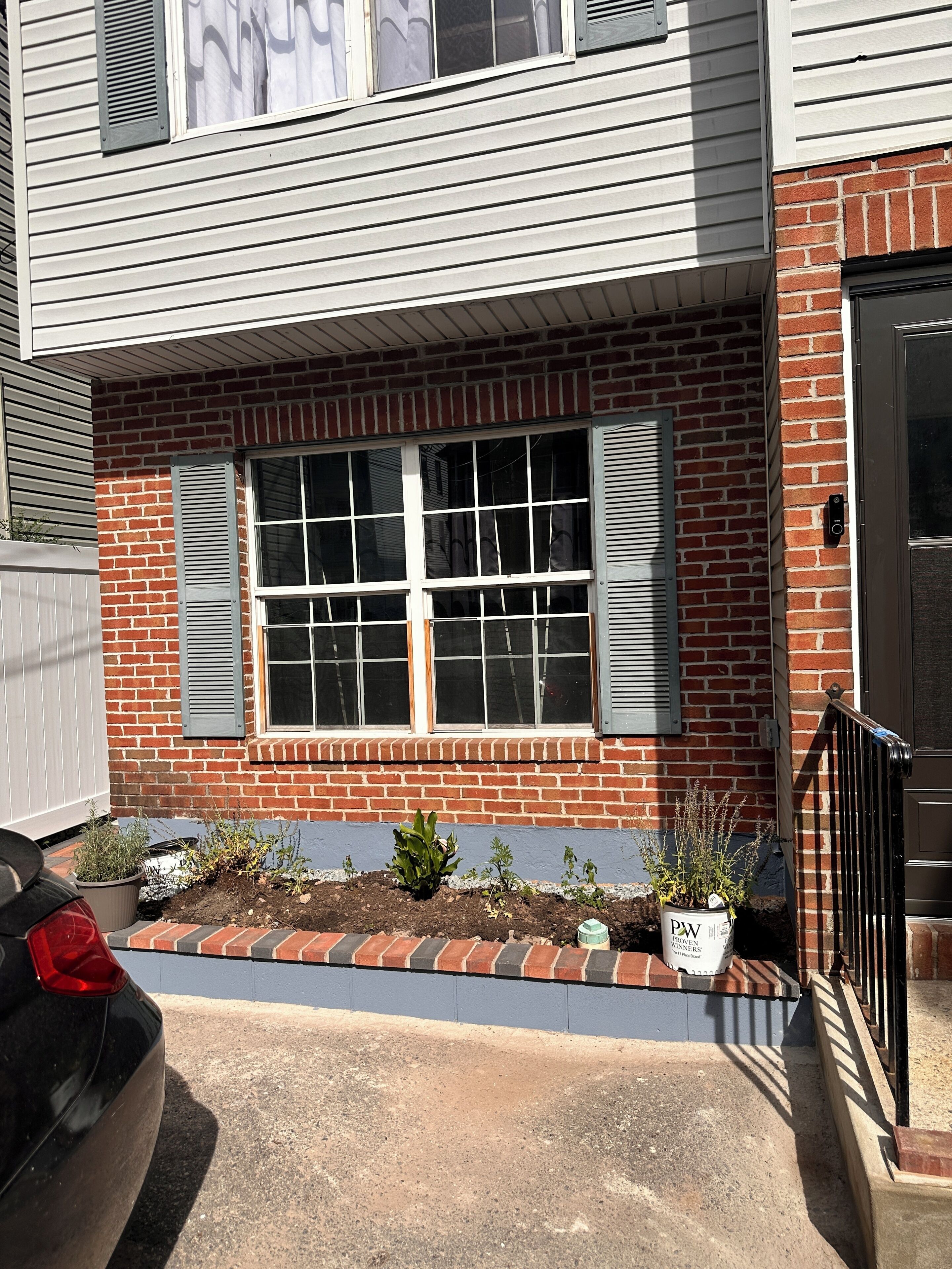 Un immeuble résidentiel à la façade en briques, doté d’une grande fenêtre avec volets, d’un petit jardin agrémenté de plantes en pot et d’une voiture noire garée devant.