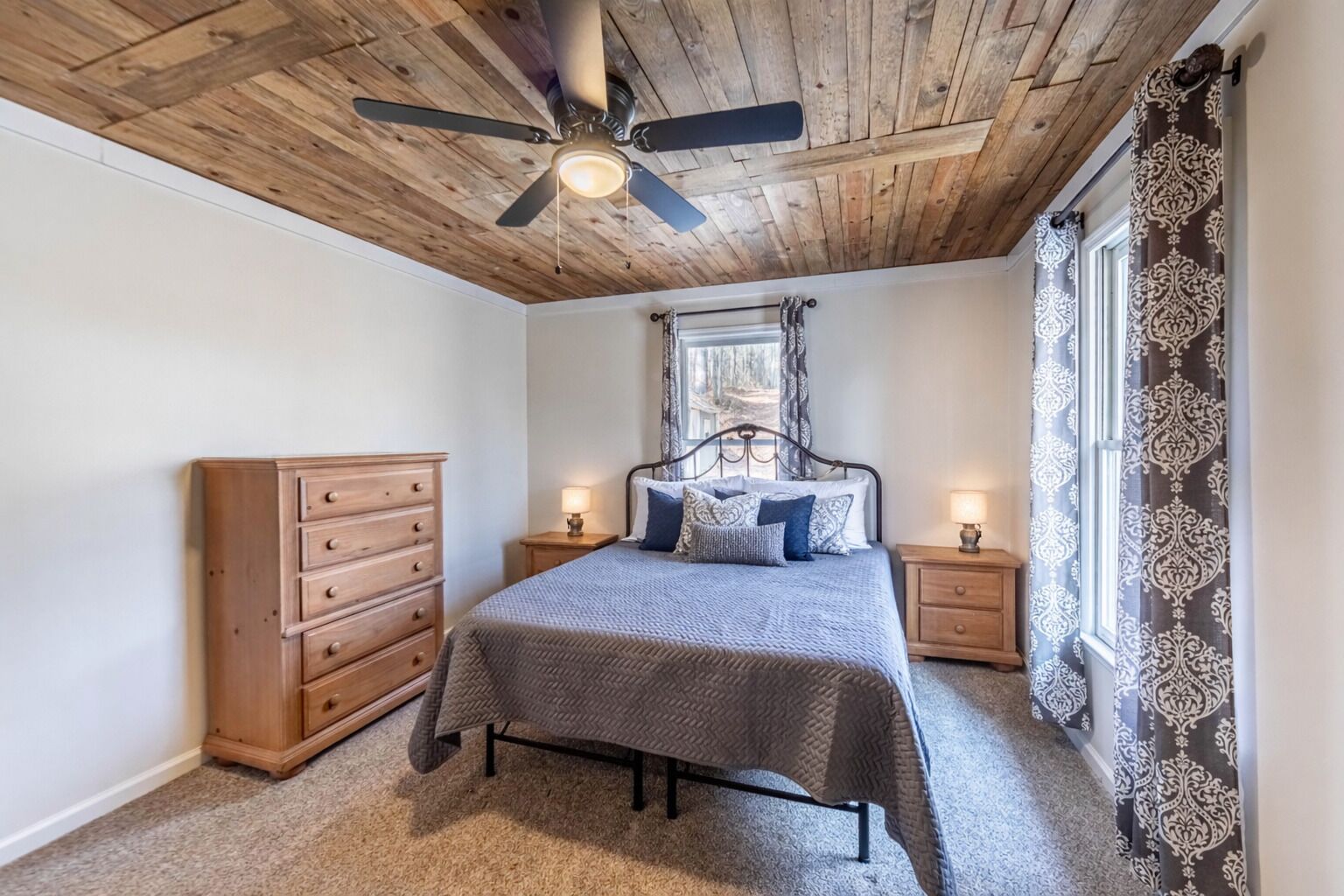 A bedroom with a wooden ceiling, a bed with a grey bedspread, two nightstands with lamps, a wooden dresser, and patterned curtains.