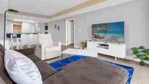 Una sala de estar moderna con un sofá, una mesa de centro y un televisor. Se observa un comedor con sillas y una mesa al fondo.