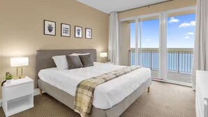 Habitación de hotel con una cama grande, ventilador de techo, balcón con vista al mar y obras de arte enmarcadas en la pared.