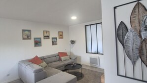 A modern living room with a grey sectional sofa, a small round coffee table, and a large window with vertical blinds. - Charming house between the countryside and the sea (SAINT NAZAIRE SUR CHARENTE)