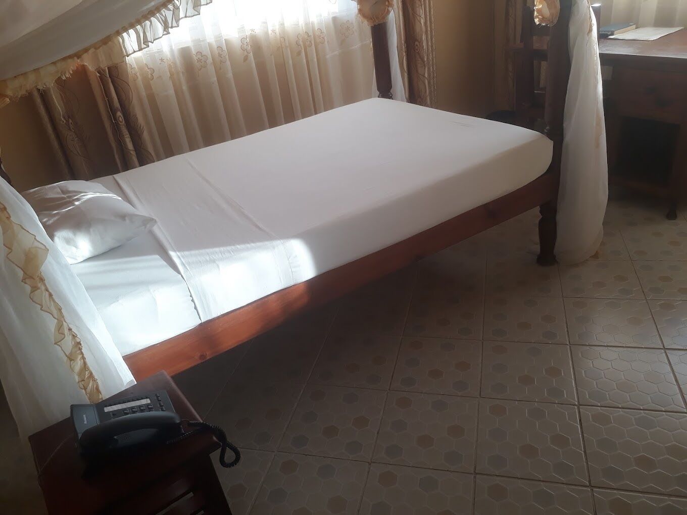 Una cama individual con colchón blanco y estructura de madera, una mesita de noche con teléfono y una cortina parcialmente abierta.