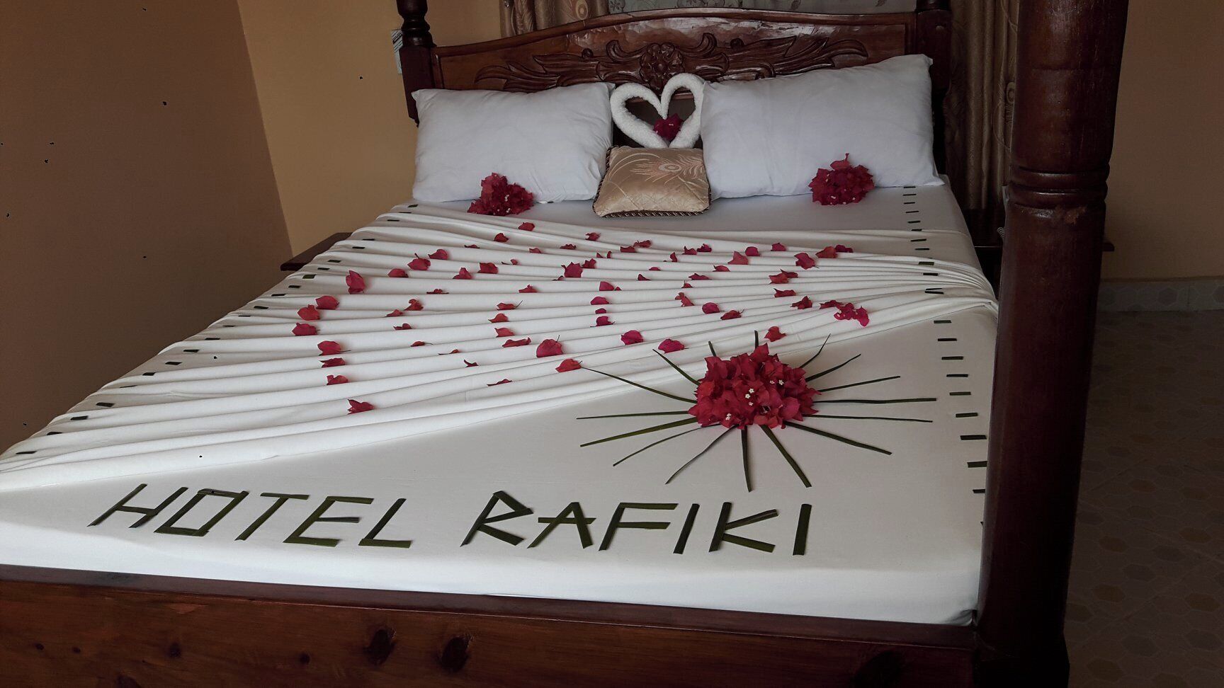 Una cama con una colcha blanca, una decoración con forma de corazón hecha de pétalos de rosa y el texto “HOTEL RAFIKI” en la colcha.