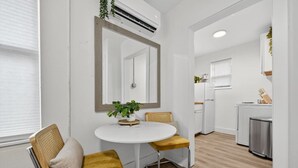 Een ruimtelijk ingerichte leefruimte met een ronde eettafel, twee stoelen, een potplant, een spiegel, een keukenhoek met een koelkast en een wasmachine.
