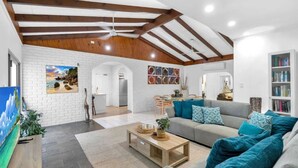Un salon spacieux avec un plafond haut, des poutres apparentes, un canapé gris agrémenté de coussins turquoise, une table basse et une bibliothèque.
