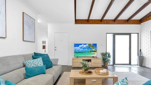 Un salon moderne avec un canapé gris, des coussins turquoise, une table basse en bois et un téléviseur à écran plat diffusant une scène de plage tropicale.