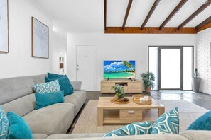 Ein modernes Wohnzimmer mit einem grauen Sofa, petrolfarbenen Kissen, einem Holztisch und einem Flachbildfernseher, der eine tropische Strandlandschaft zeigt.