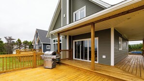Een terras met een barbecue en tuinmeubilair, een overdekte veranda en een huis met grote ramen en een houten terras.