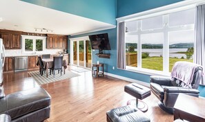 A spacious living room with a large window offering a view of a lake and mountains. - Spacious Home with Stunning Ocean Bay Views (Placentia)