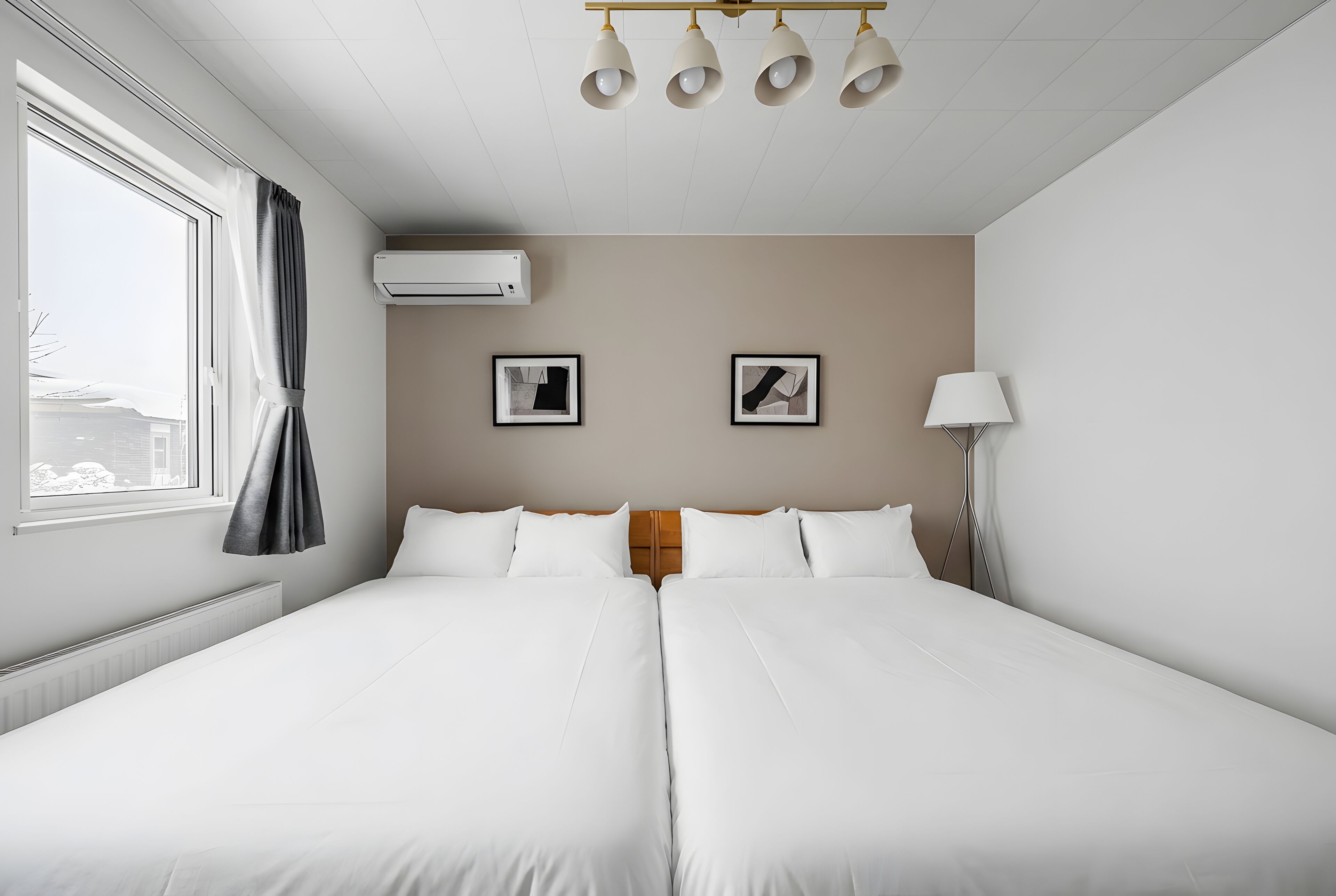 A hotel room with two beds, a window with curtains, an air conditioning unit, and two framed pictures on the wall.
