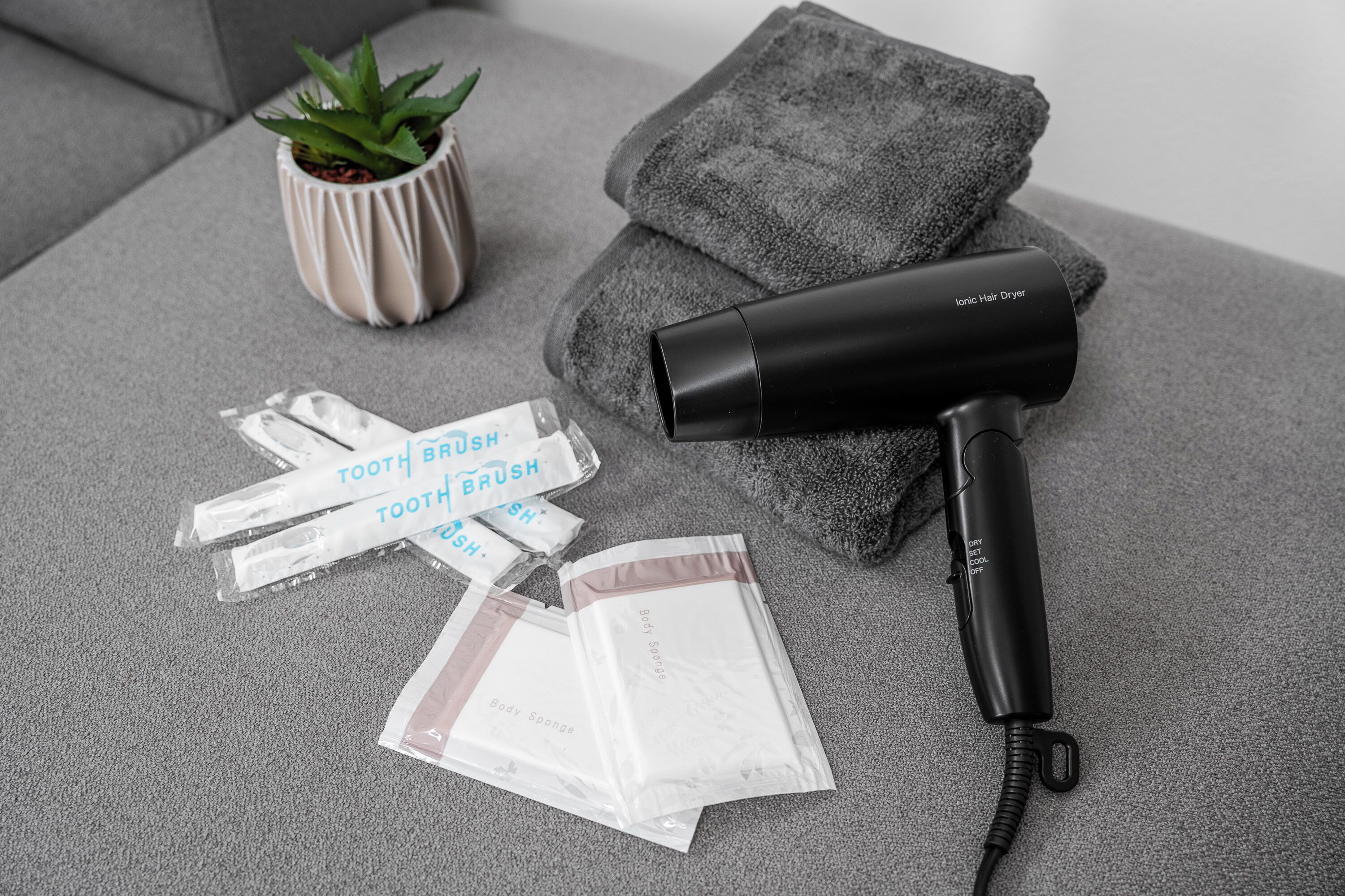 Un sèche-cheveux, une serviette et des produits d’hygiène personnelle emballés, posés sur une surface grise.