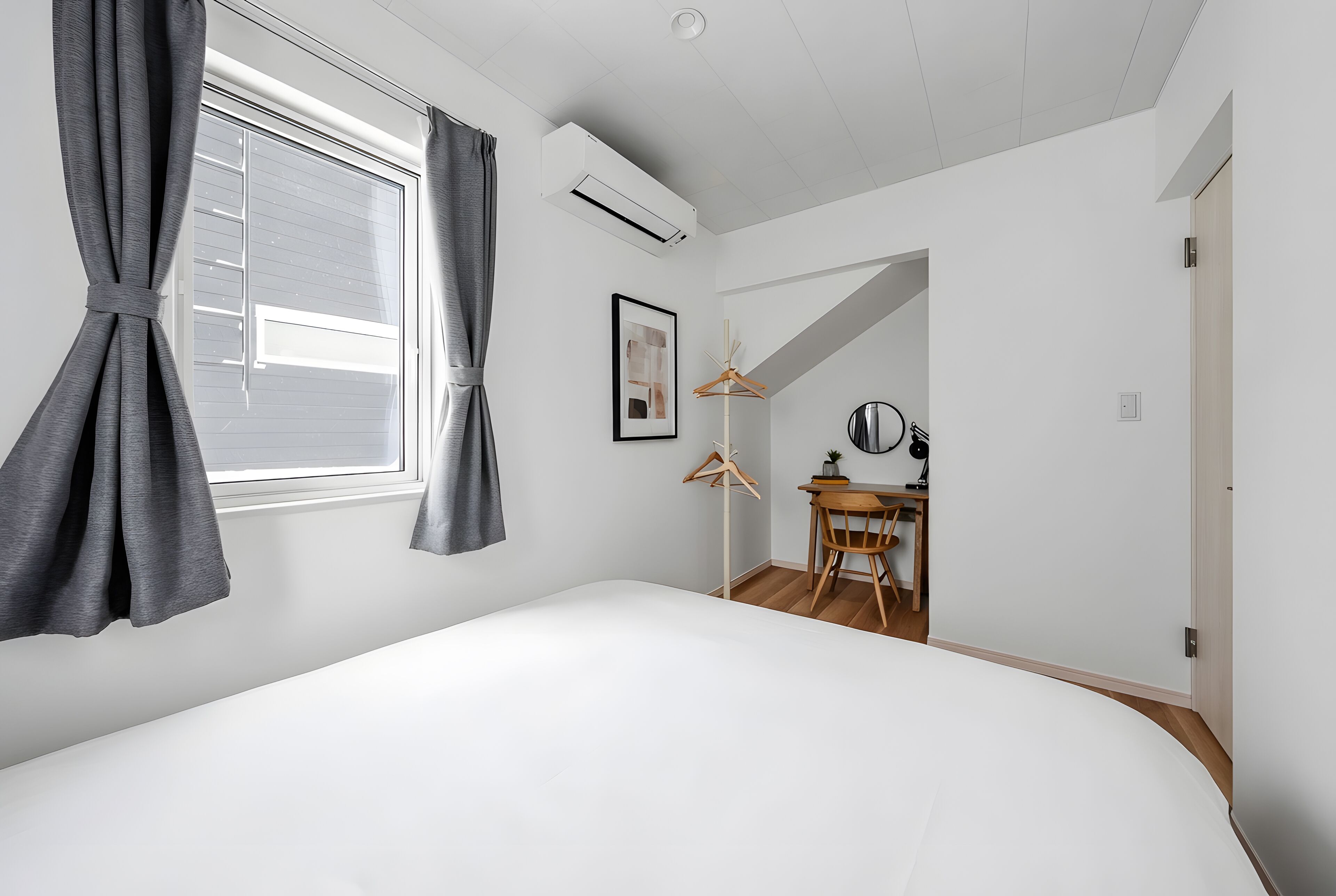 A bedroom with a large bed, a window with curtains, an air conditioning unit, a desk with a chair, and a coat rack.