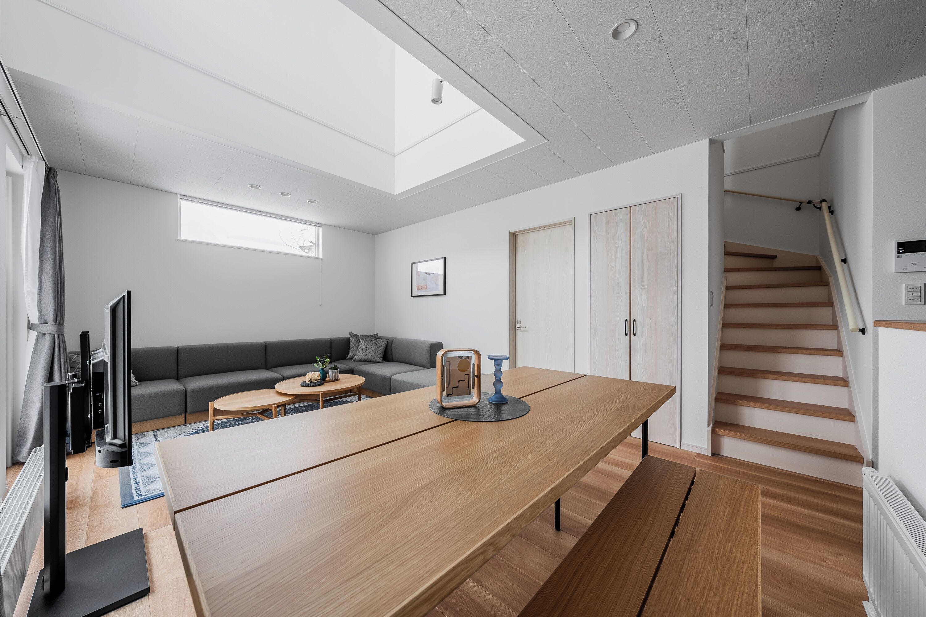 A modern living room with a large wooden table, a grey sofa, and a staircase leading to an upper level.