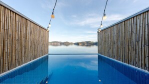 Piscina amb parets de bambú i llums penjants, cel blau i vista llunyana de les colines.