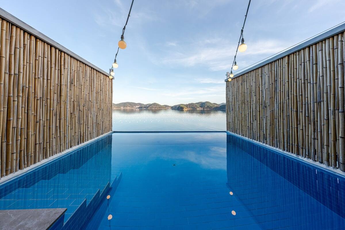 Una piscina con paredes de bambú e iluminación colgante, un cielo despejado y una vista distante de colinas.
