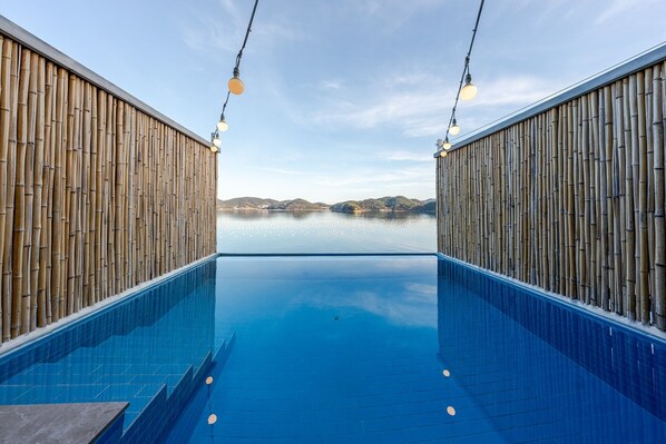 A pool with bamboo walls and hanging lights, a clear sky, and a distant view of hills. - Geoje Rainbow Kids Pool Villa (Geoje)