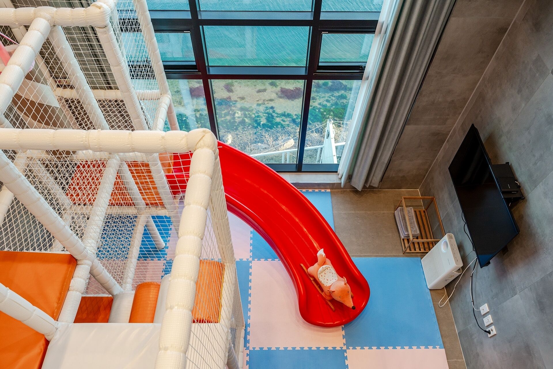 Un espace de jeux pour enfants avec un toboggan rouge, un filet d’escalade blanc et une vue dégagée grâce à de grandes fenêtres.
