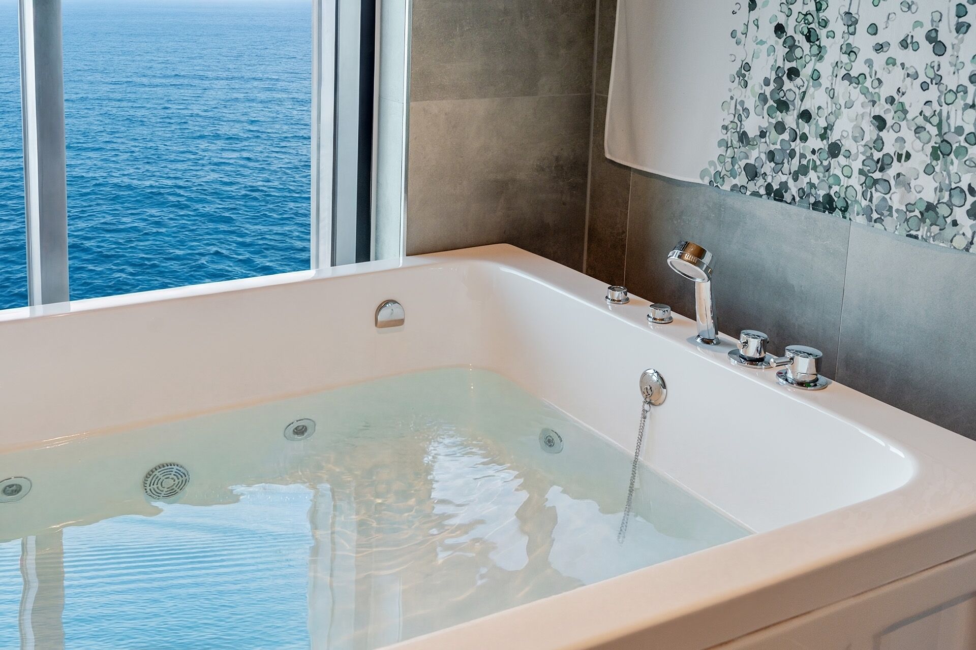 A modern bathroom with a bathtub, a window with a view of the sea, and a wall with a decorative pattern.