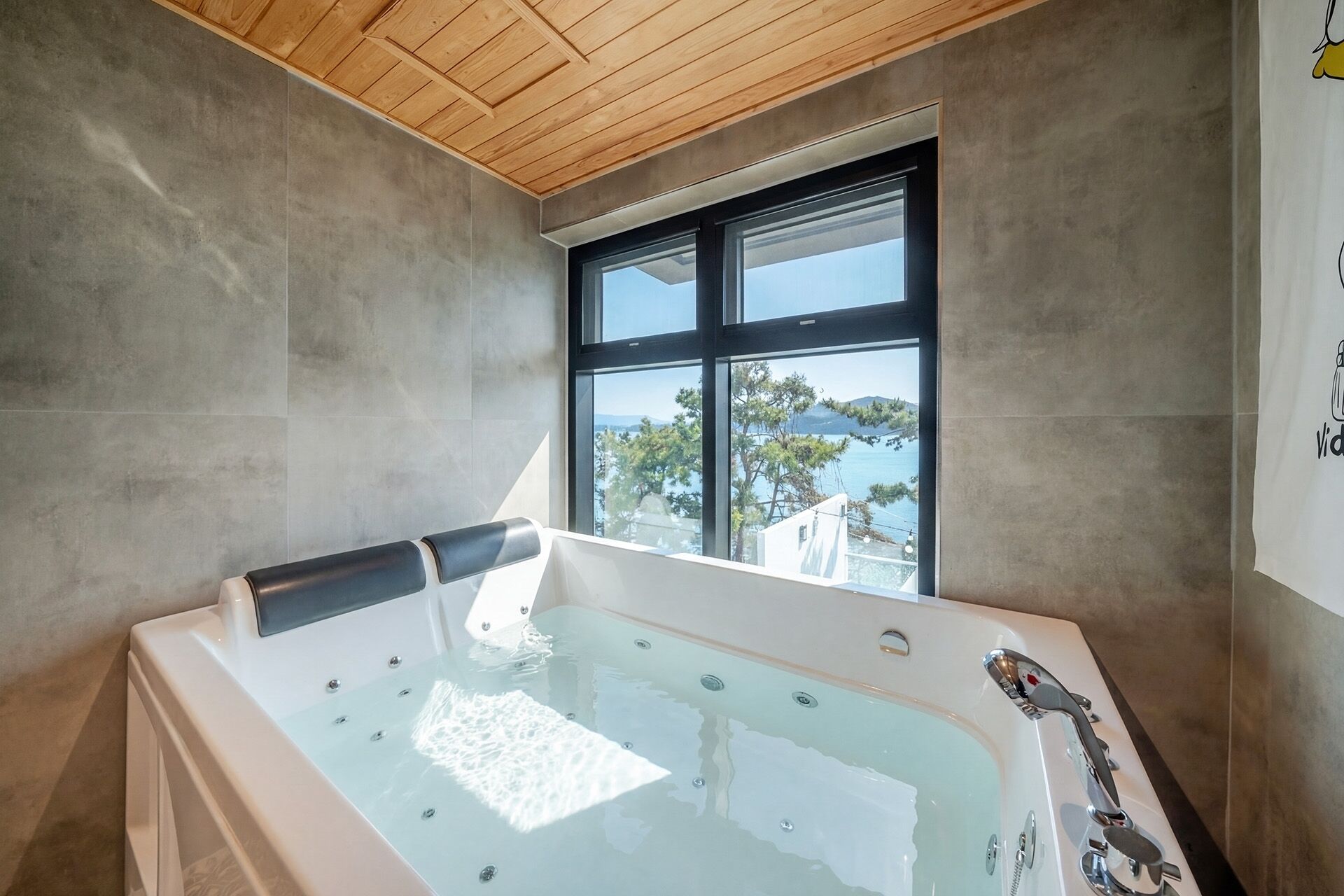 A modern bathroom with a large bathtub, a wooden ceiling, and a view of a body of water through a window.