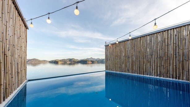 A swimming pool with bamboo walls and string lights, surrounded by a calm body of water and distant hills. - Geoje Rainbow Kids Pool Villa (Geoje)
