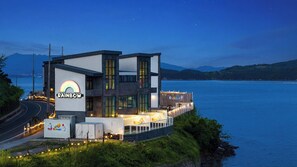 A modern building with a 'RAINBOW' sign, located by a body of water with mountains in the background. - Geoje Rainbow Kids Pool Villa (Geoje)
