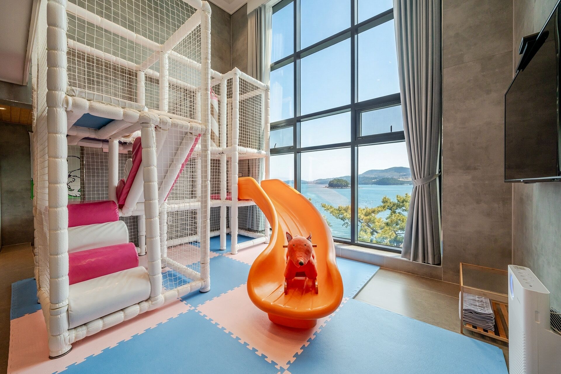 Un espace de jeux pour enfants avec un toboggan et un filet d’escalade, de grandes fenêtres offrant une vue sur la mer et un aménagement intérieur moderne.