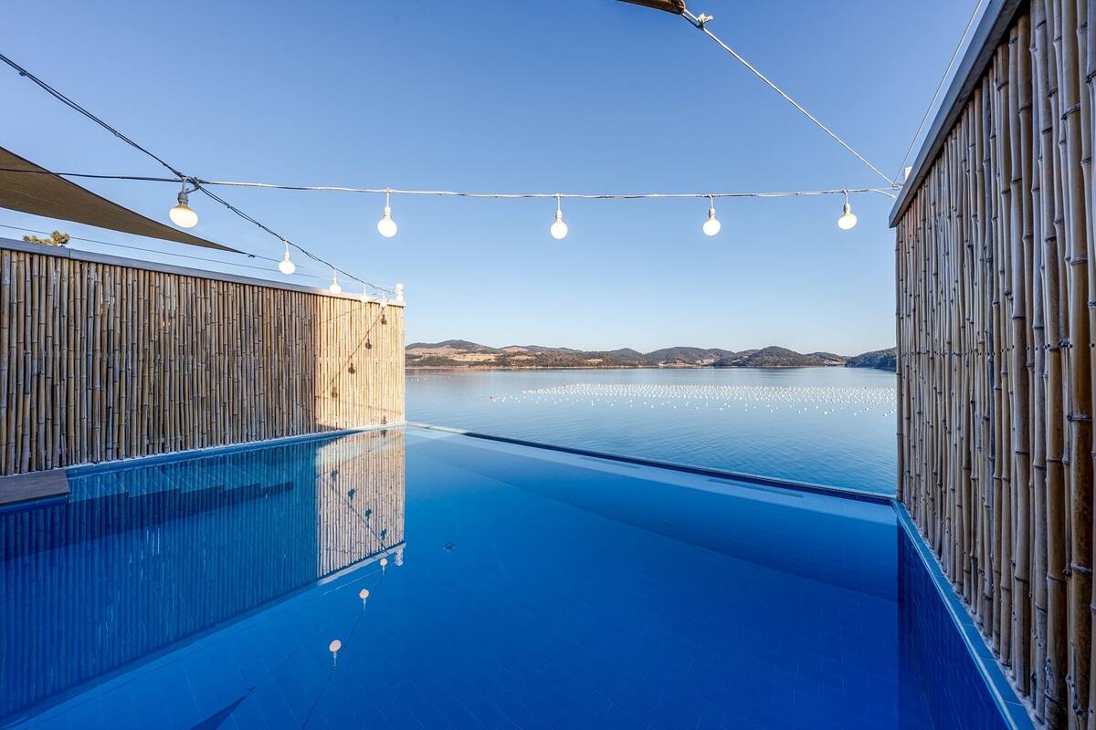 Piscina azul cristalina con paredes de bambú, luces colgantes y vistas a colinas distantes.