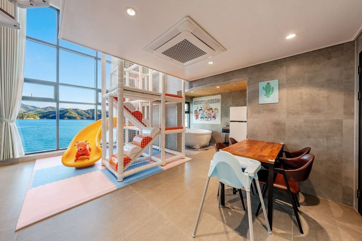 Una habitación con área de juegos, una mesa de comedor con sillas, una bañera y vista al agua.