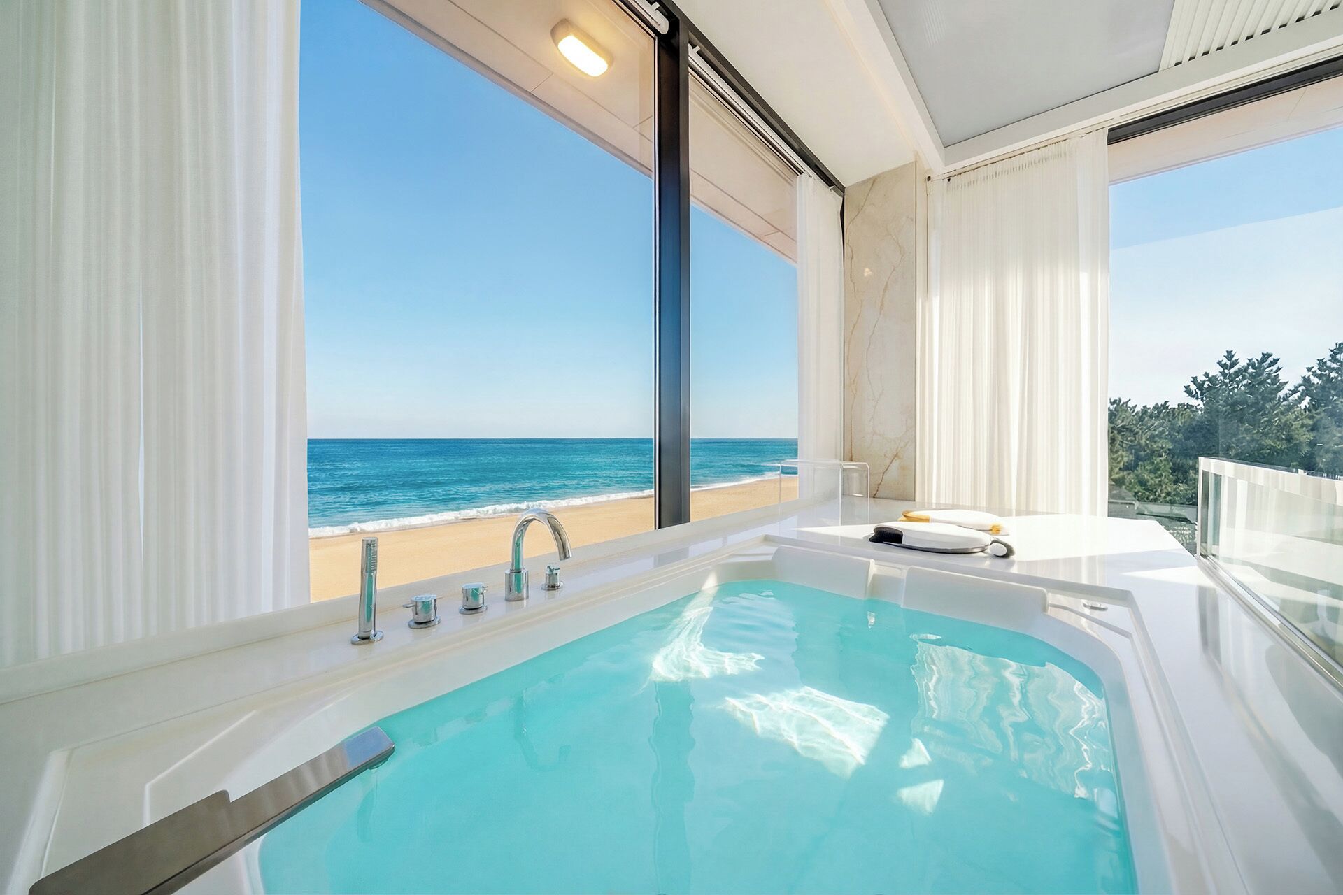A modern bathroom with a large bathtub, a shower, and a view of the beach.