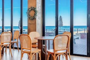 A beachfront restaurant with wicker chairs and a wooden table, large windows, and a wreath on a pillar.