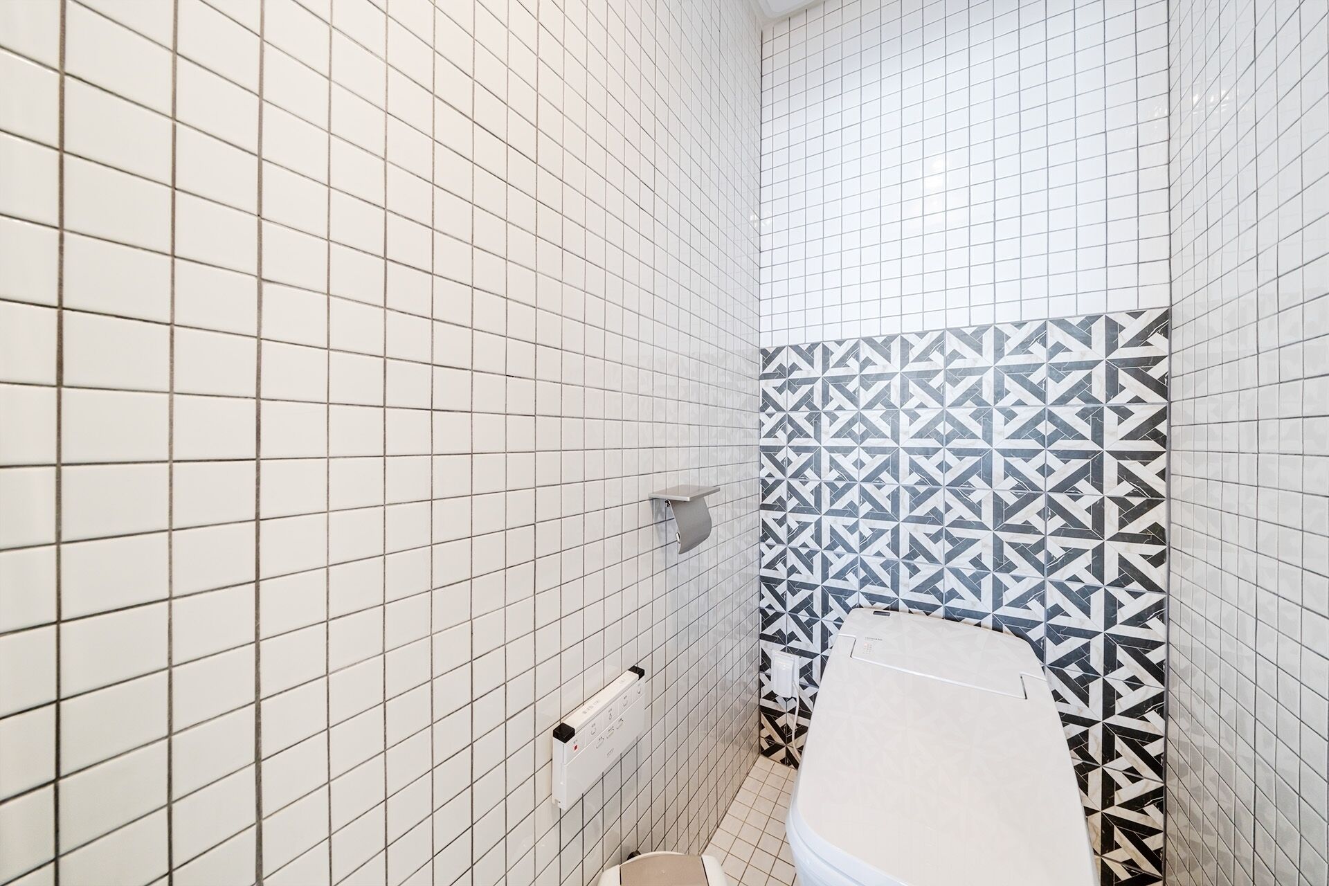 A bathroom with white tiled walls, a patterned tile accent wall, a toilet, and a towel rack.