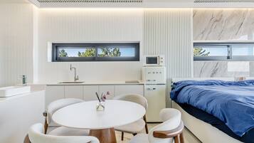 A modern bedroom with a bed, a small kitchenette, a round dining table with chairs, and a window with a view of trees.