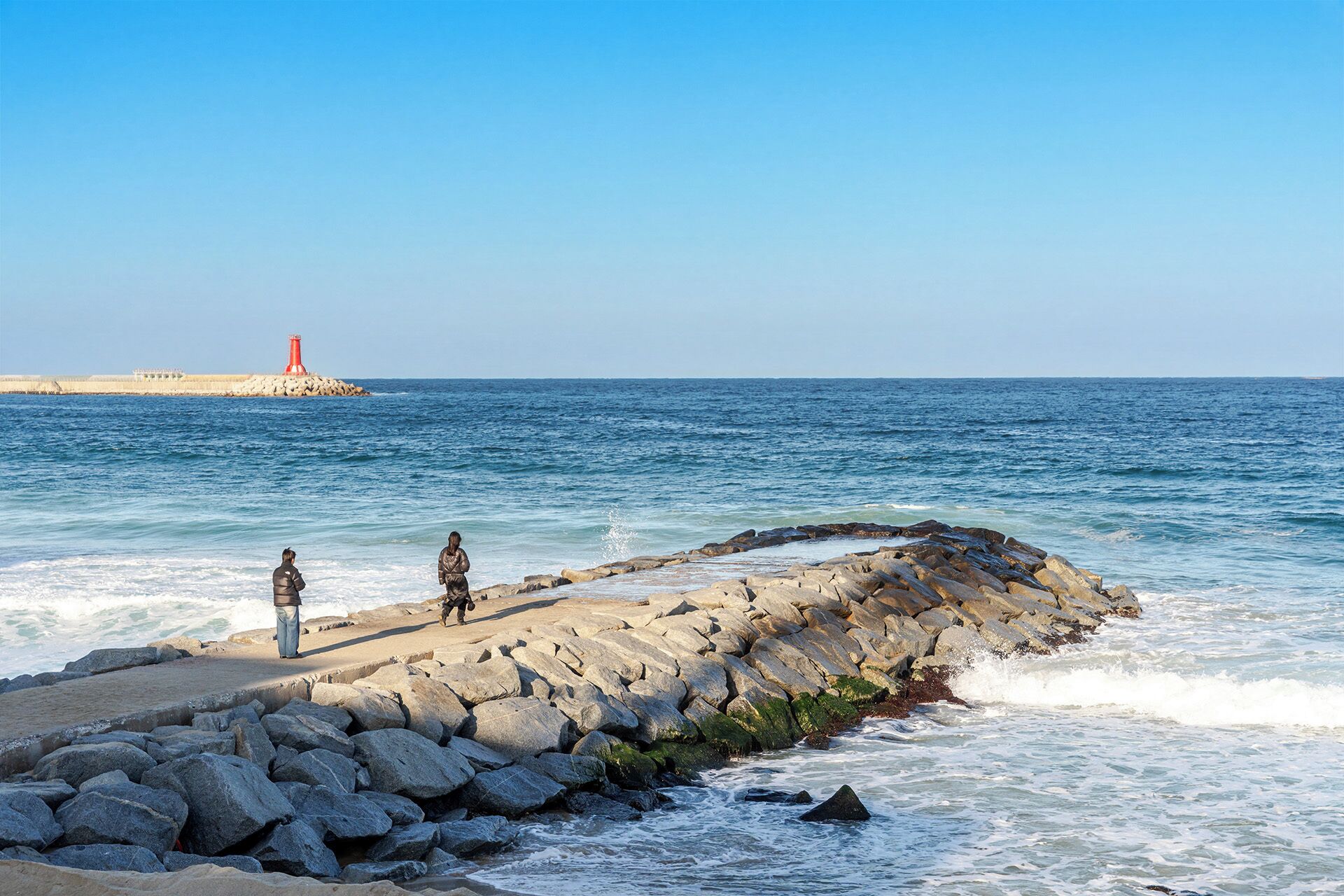 Una costa rocciosa con le onde che si infrangono, un faro in lontananza e due persone in piedi sugli scogli.