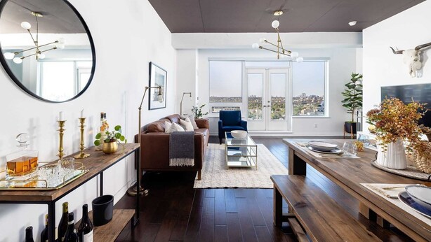 Een moderne woonkamer met een grote spiegel, een bruine leren bank, een blauwe fauteuil en een eethoek met een houten tafel en bankje.