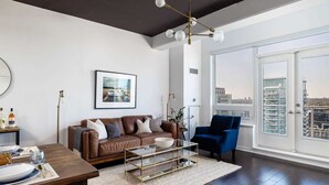 Een moderne woonkamer met een bruine leren bank, een blauwe fauteuil, een glazen salontafel en een stadsgezicht door grote ramen.