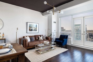 A modern living room with a brown leather sofa, a blue armchair, a glass coffee table, and a city view through large windows.