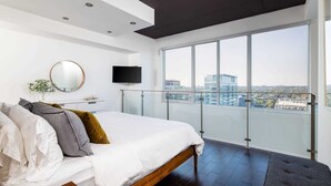 Een moderne slaapkamer met een groot bed, een televisie, een balkon met uitzicht op de stad, en een ronde spiegel aan de muur.