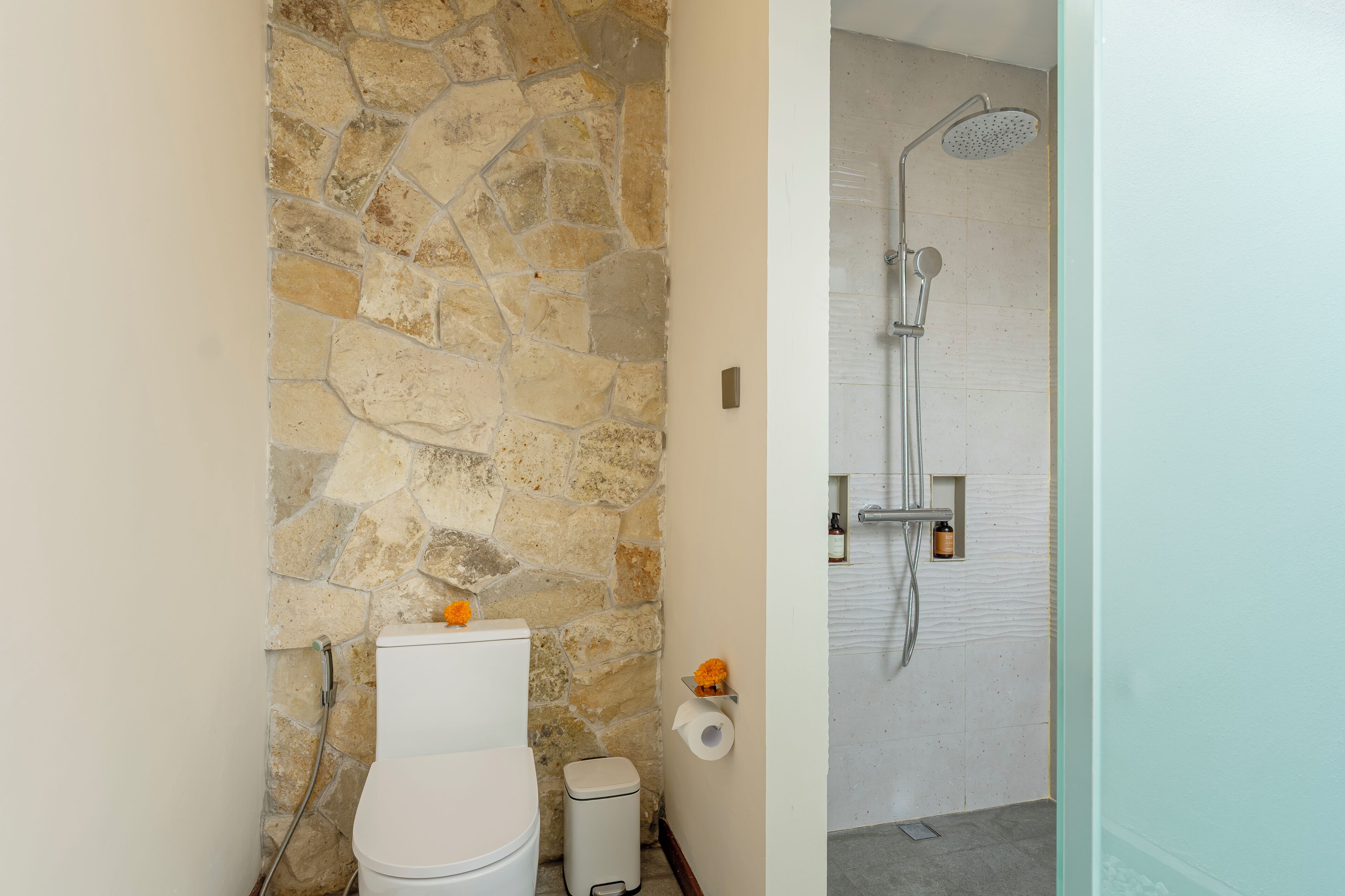 A bathroom with a stone wall, a toilet, a trash bin, a shower with a handheld showerhead, and a toilet paper holder with a flower.