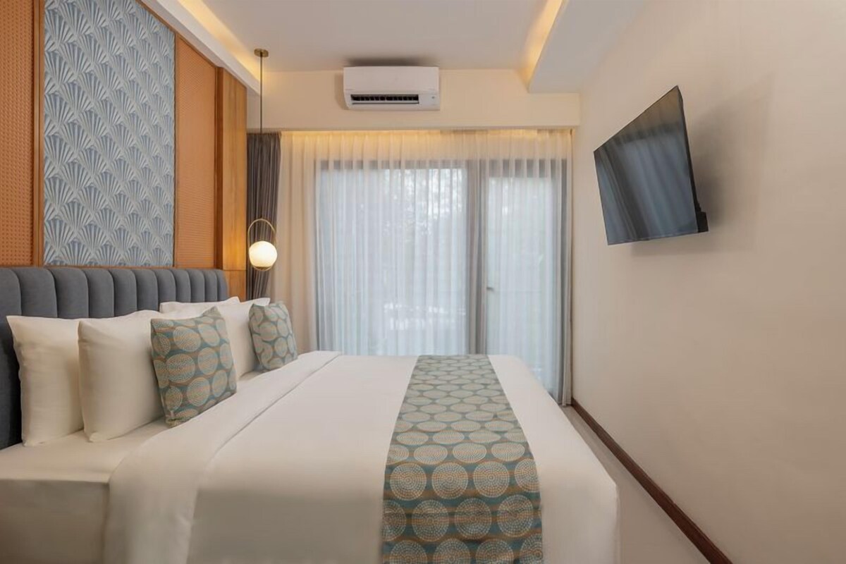A hotel room with a bed, a flat-screen TV mounted on the wall, and a window with curtains.