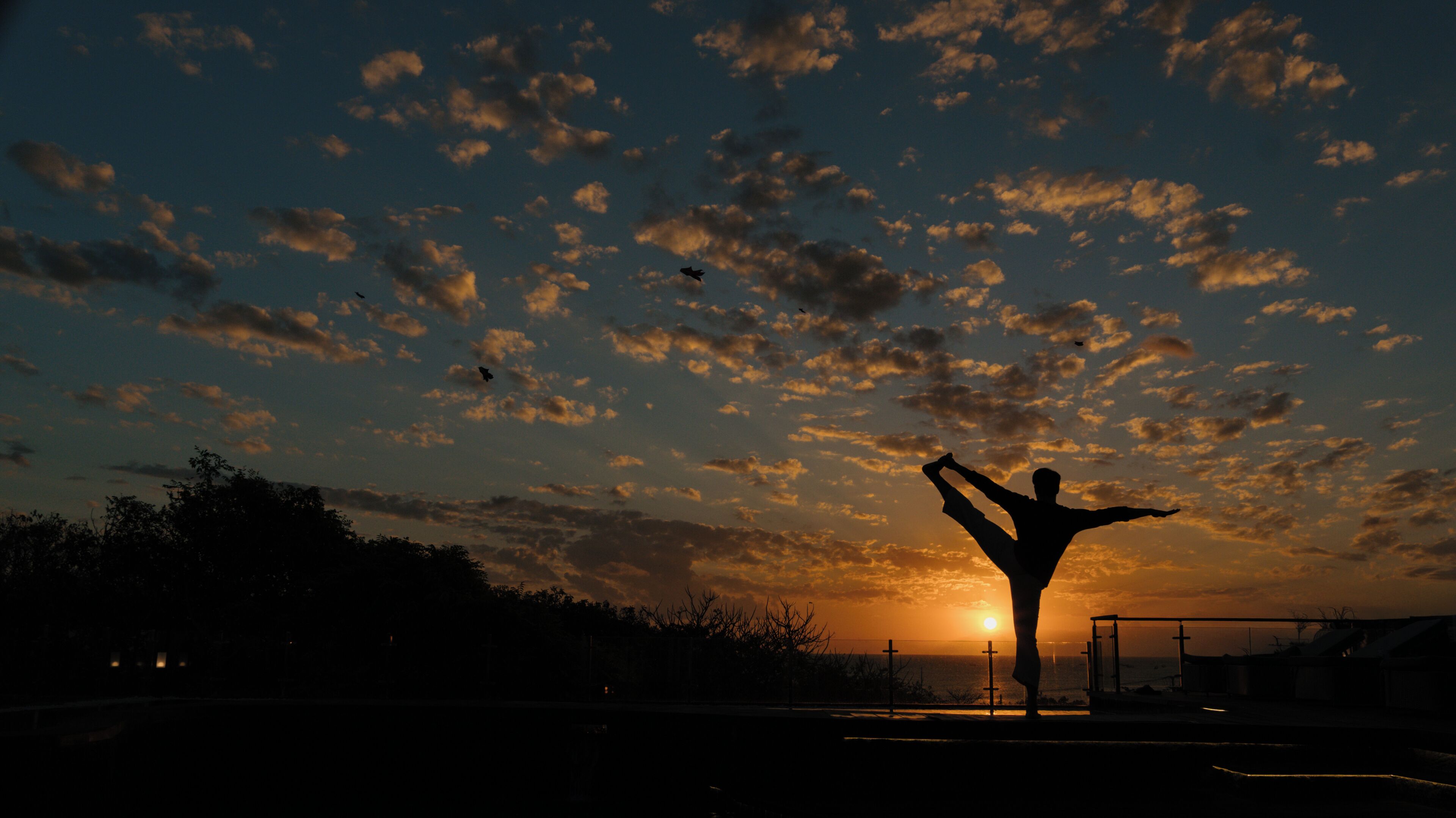 A silhouette of a person performing a yoga pose against a sunset sky with clouds.