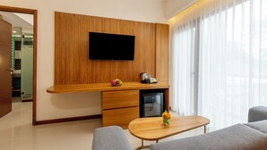 Egy modern nappali, melyben egy síkképernyős televízió van felszerelve egy fa borítású falra, egy kis hűtőszekrény, egy fa kávézóasztal és szürke színű kanapék találhatók.