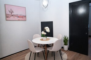 Un coin repas avec une table ronde, quatre chaises, un vase de fleurs blanches, un tableau encadré au mur et une plante en pot.