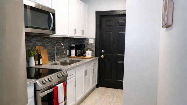 A compact kitchen with white cabinets, a stainless steel microwave, a sink, and a black door. - Fully Furnished One-Bedroom Near Downtown & Medical Center (Houston)