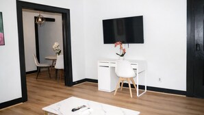 A modern living room with a white desk, a flat-screen TV mounted on the wall, a dining area with a table and chairs, and a hallway leading to another room. - Fully Furnished One-Bedroom Near Downtown & Medical Center (Houston)