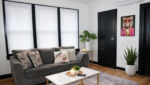 A living room with a grey sofa, patterned cushions, a small white table with a remote control, a potted plant, and a painting of a tiger on the wall. - Fully Furnished One-Bedroom Near Downtown & Medical Center (Houston)