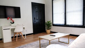 A modern living room with a white sofa, a coffee table, a desk with a chair, a potted plant, and a black door. - Fully Furnished One-Bedroom Near Downtown & Medical Center (Houston)