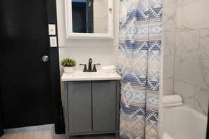 Une salle de bain avec une porte noire, un lavabo blanc avec un robinet noir, une petite plante en pot et un rideau de douche à motifs.